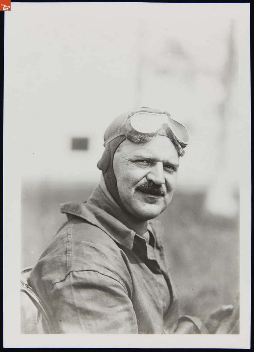 Man with broom mustache in jacket and soft racing helmet, with goggles pushed up on forehead