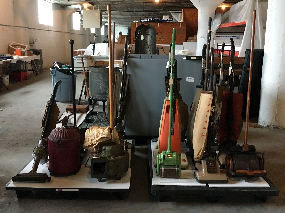 A group of vacuum cleaners sits on two pallets in a large room with other boxes, worktables, and items in the background