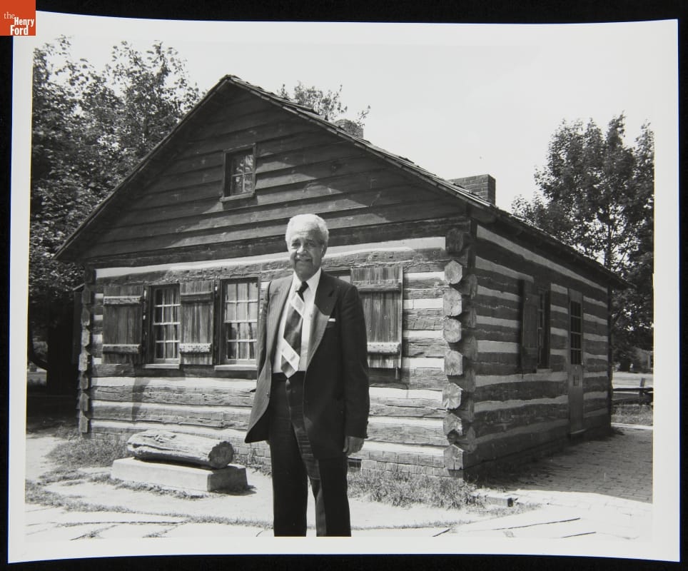 Black-and-white photo of man in suit standing in front of small wooden cabin