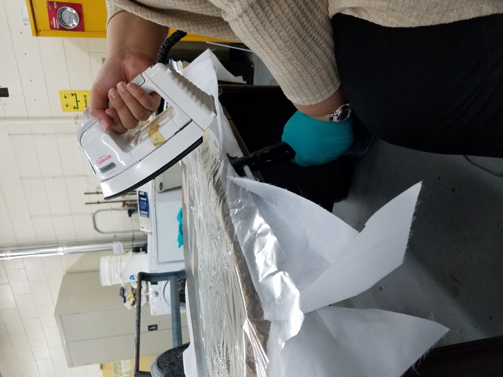 Person wearing gloves holds an iron to a flat item on a table in a lab