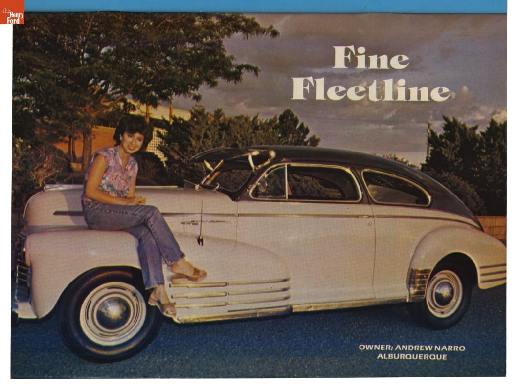 Woman sits on fender of large, bulbous white car with gray top