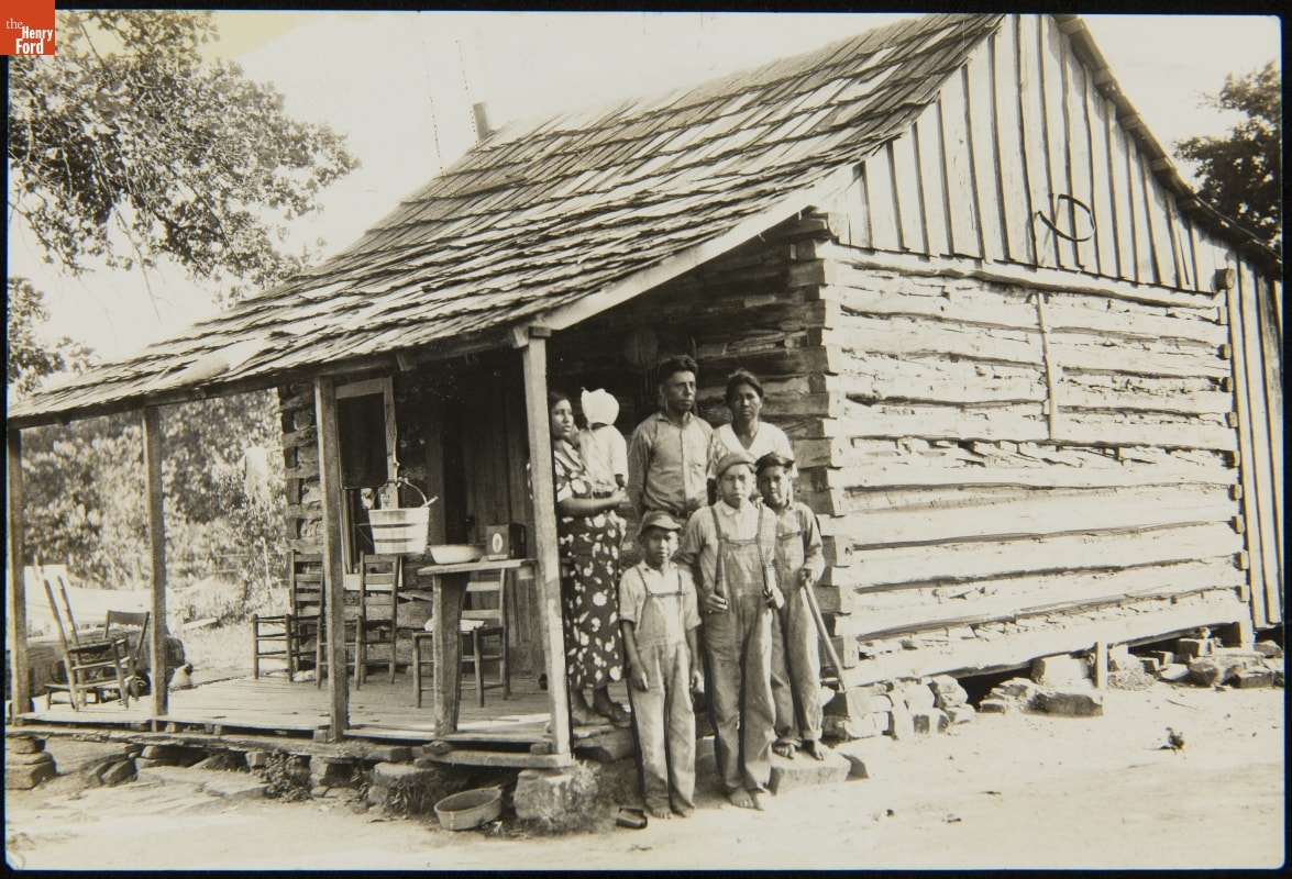 Black-and-white photo of group of adults and children standing on or near porch of very minimalistic wooden house