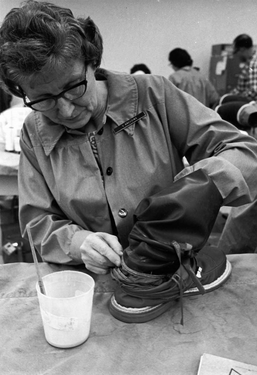 Woman in a smock with nametag works with her hand inside a boot; a cup with a brush in it sits nearby