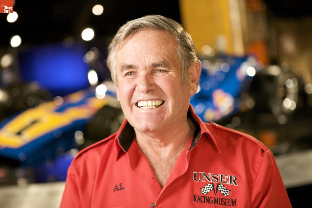 Man in red shirt with text and checkered flag logo smiles at camera; out-of-focus race car in background