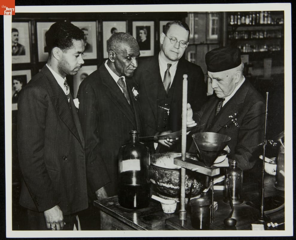 Black-and-white image of four men in suits looking at a piece of equipment on a table in a  workroom or laboratory
