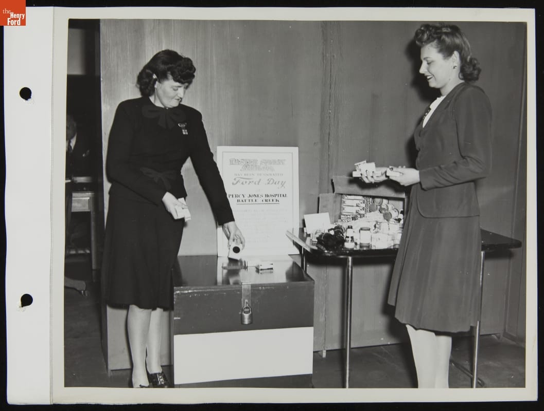 Collecting Gifts at Ford Rotunda for Percy Jones Veterans Hospital in Battle Creek, Michigan, April 1944 Two women with small items in their hands stand by a table and box with additional items and a sign