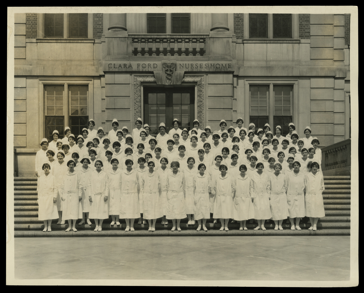 Group of women in nurses' uniforms stand on the steps of a building inscribed "Clara Ford Nurses Home"