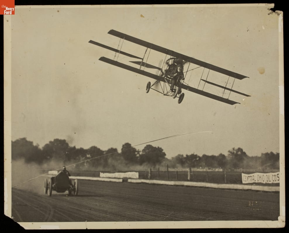 Early airplane flies low above a race car on a dirt track