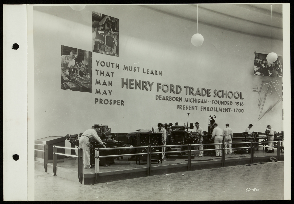Boys/young men work at a variety of machinery under a wall containing images and text