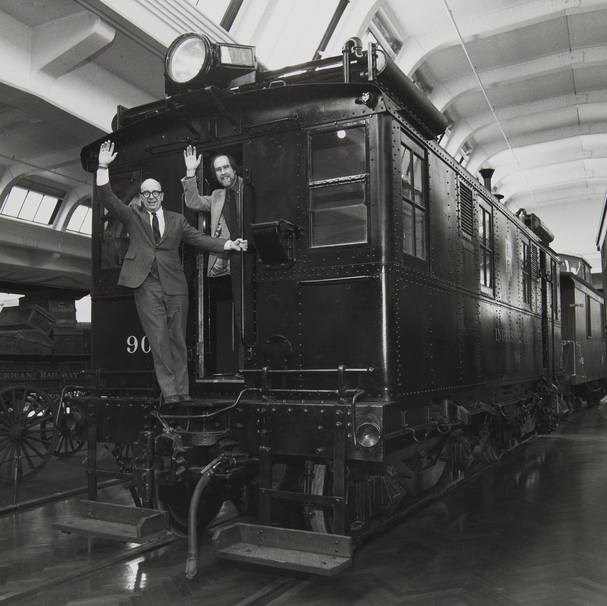 Two men wave from the back of a rail car on tracks in a large room