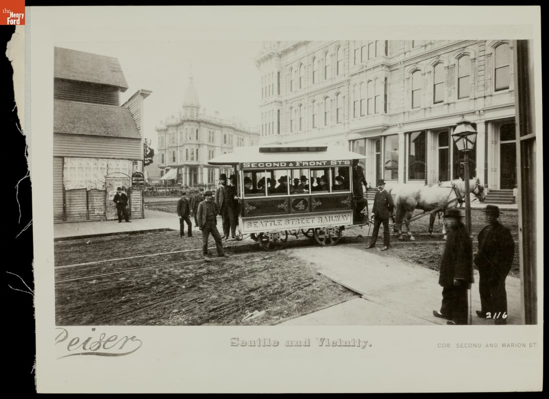 Street scene with a horse-drawn streetcar in middle of road and buildings and pedestrians along the streets