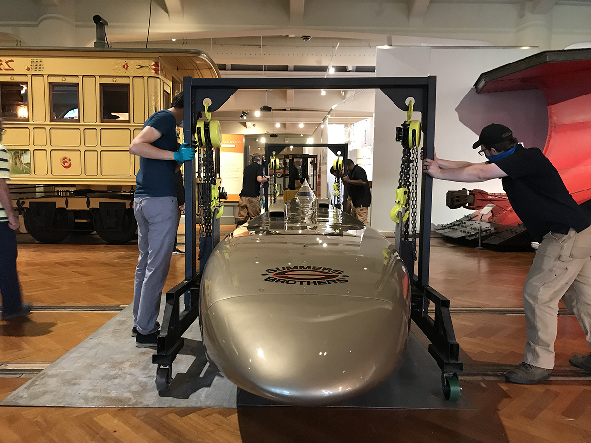 Men guide a golden car suspended from crane-like structures past railcars and streetcars