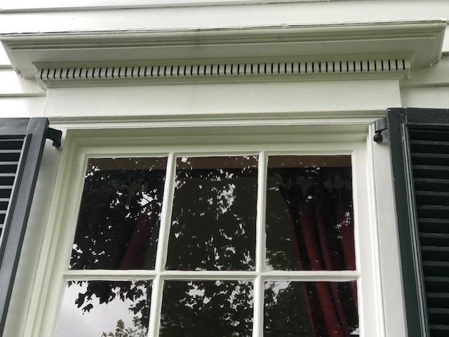 Top of a window, showing decorative trim above, and part of black shutters to either side; maroon curtains visible inside