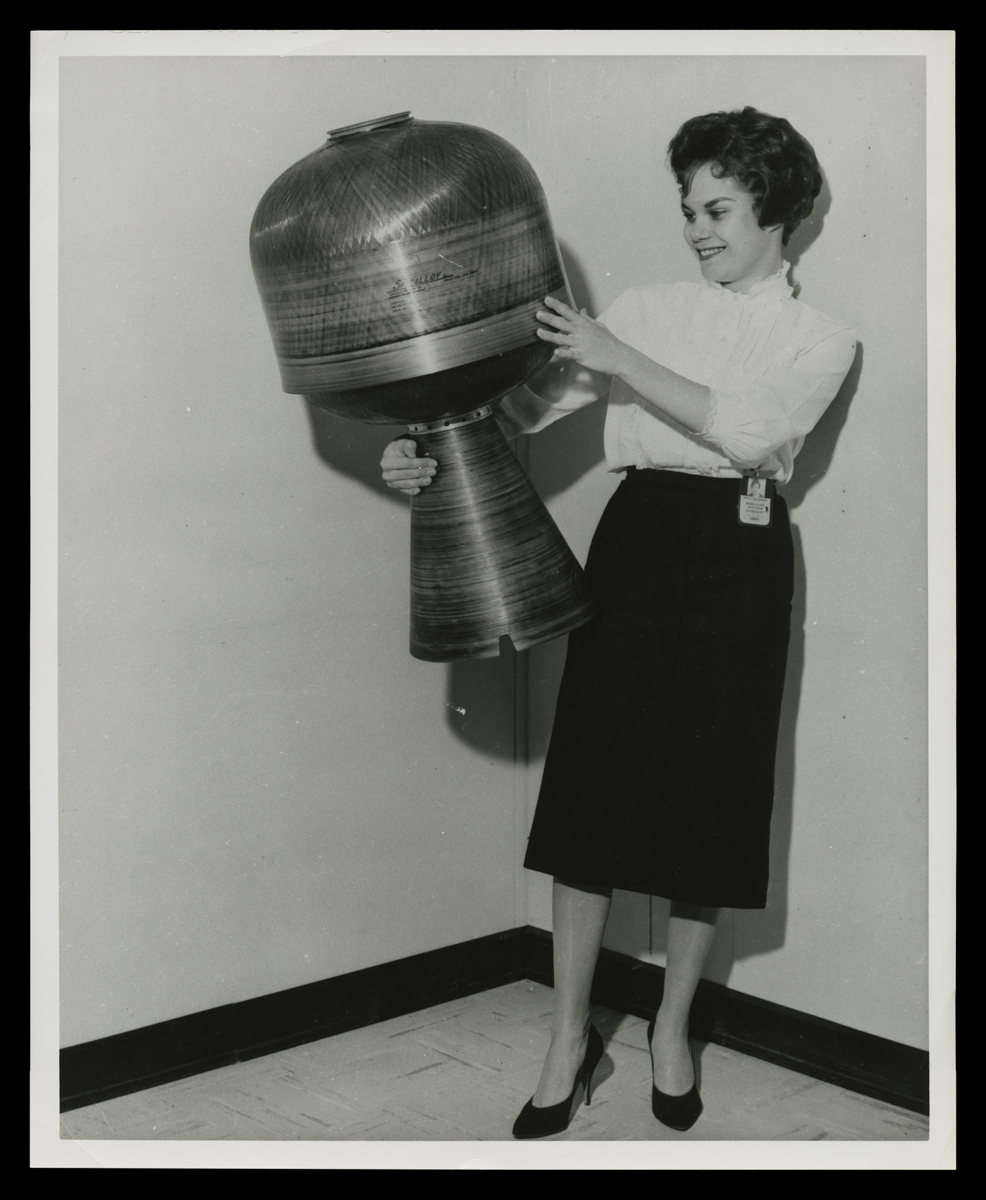 Black-and-white photo of smiling woman holding contraption with cone-shaped bottom section and dome-shaped top section 