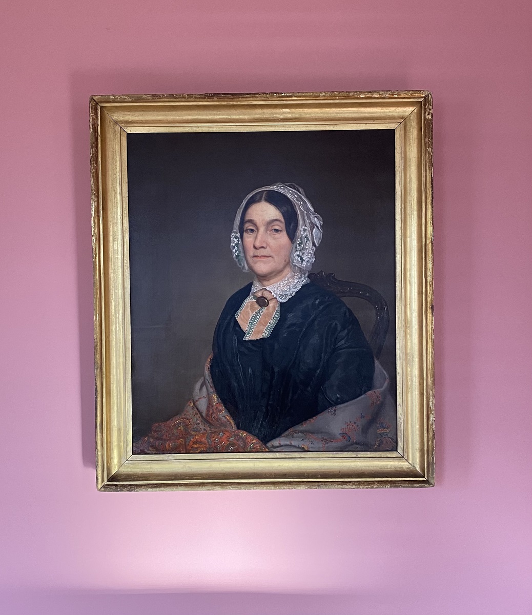 Gold-framed portrait of woman in black dress, shawl, and white lace bonnet, hanging on pink wall