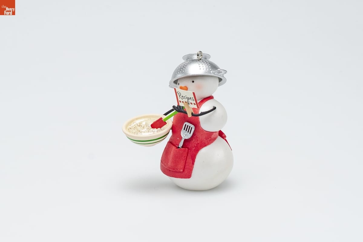 Ornament of a snowman wearing a red apron with a spatula in the pocket and a colander on its head, holding a bowl of dough in one hand and a card labeled "Recipes" in the other