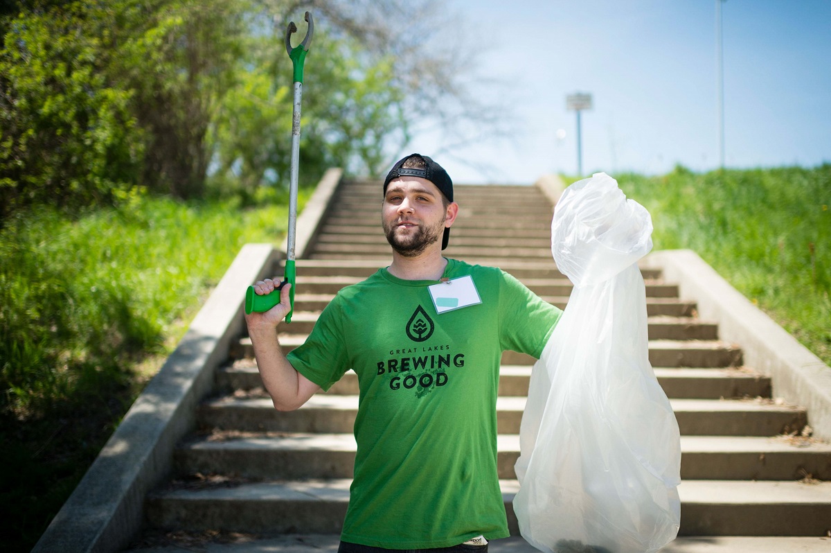 Man in baseball cap and green t-shirt holds up grabber and trash bag while standing on concrete steps with trees and grass on either side