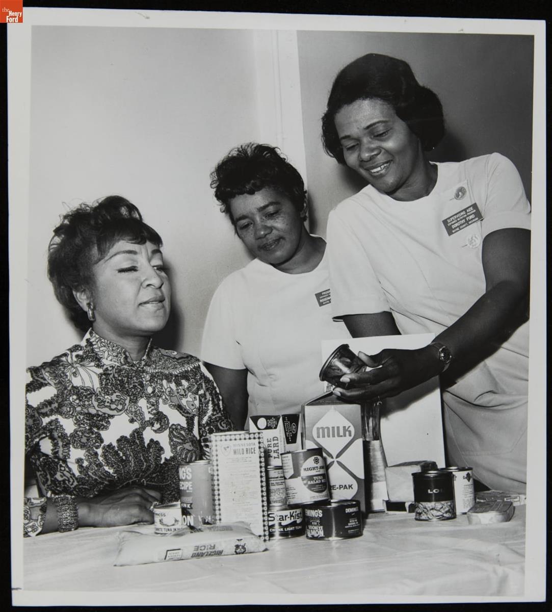 Three women of color standing and sitting around a table containing canned, boxed, and bagged food items