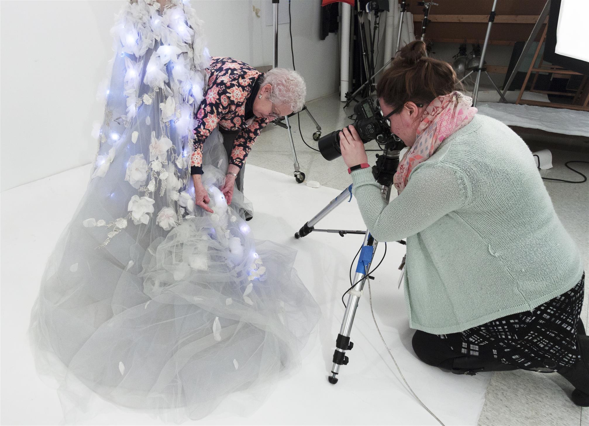 Conservator Fran Faile holds up a detail on the Cognitive Dress as Jillian Ferraiuolo photographs it. Woman in foreground looks through camera as a second woman holds up part of a dress skirt