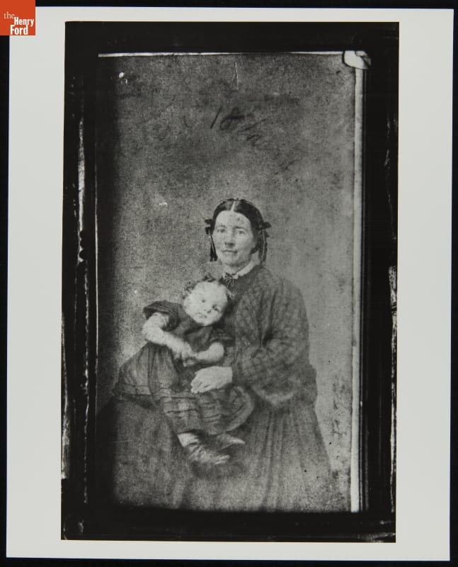 Framed portrait of woman in dress with hair in two long coils on either side of her face, holding a young child