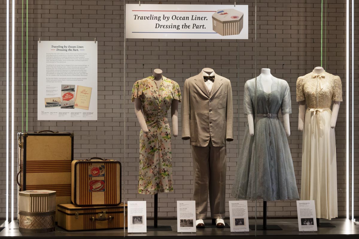 Large museum display case containing four mannequins wearing clothing, luggage, and signage