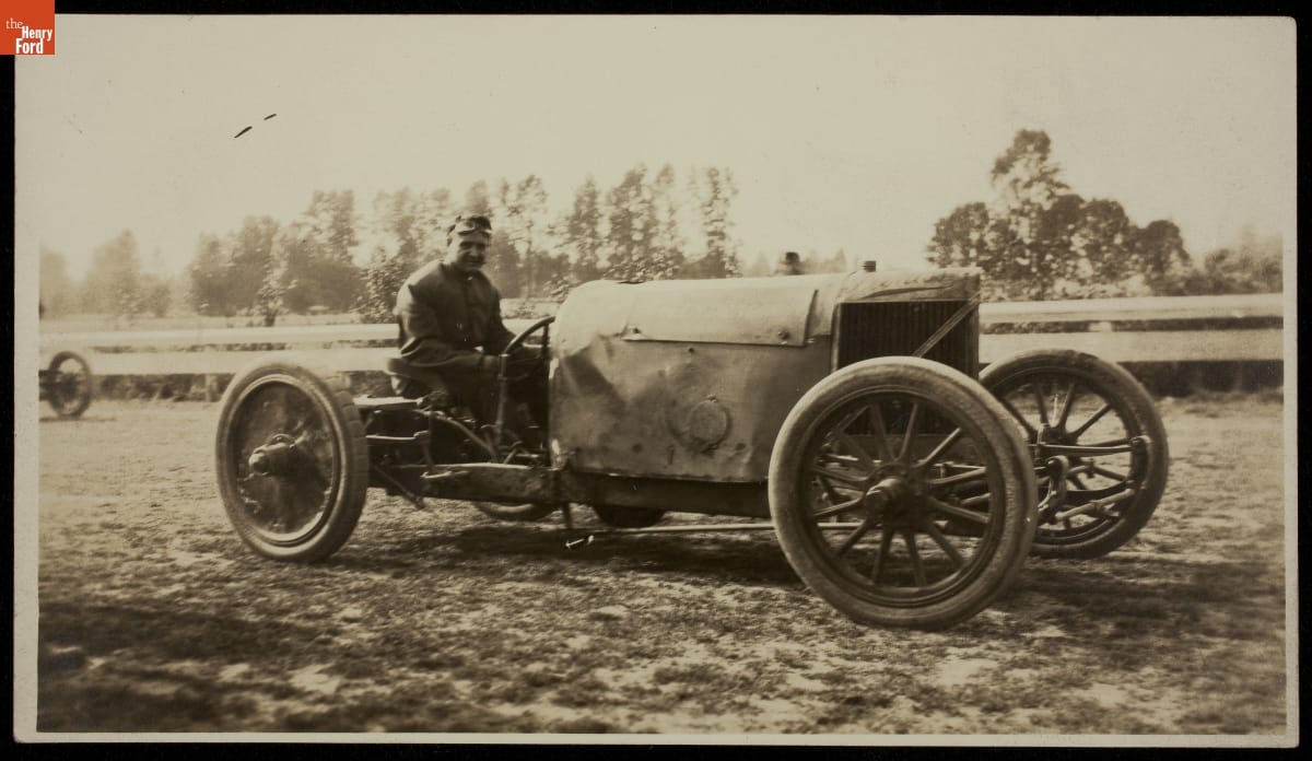 Man sits behind wheel of early open race car on a dirt track