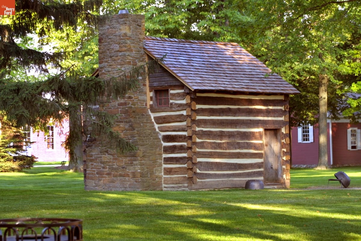 Small log cabin with shingled roof and stone fireplace, surrounded by grass and other buildings