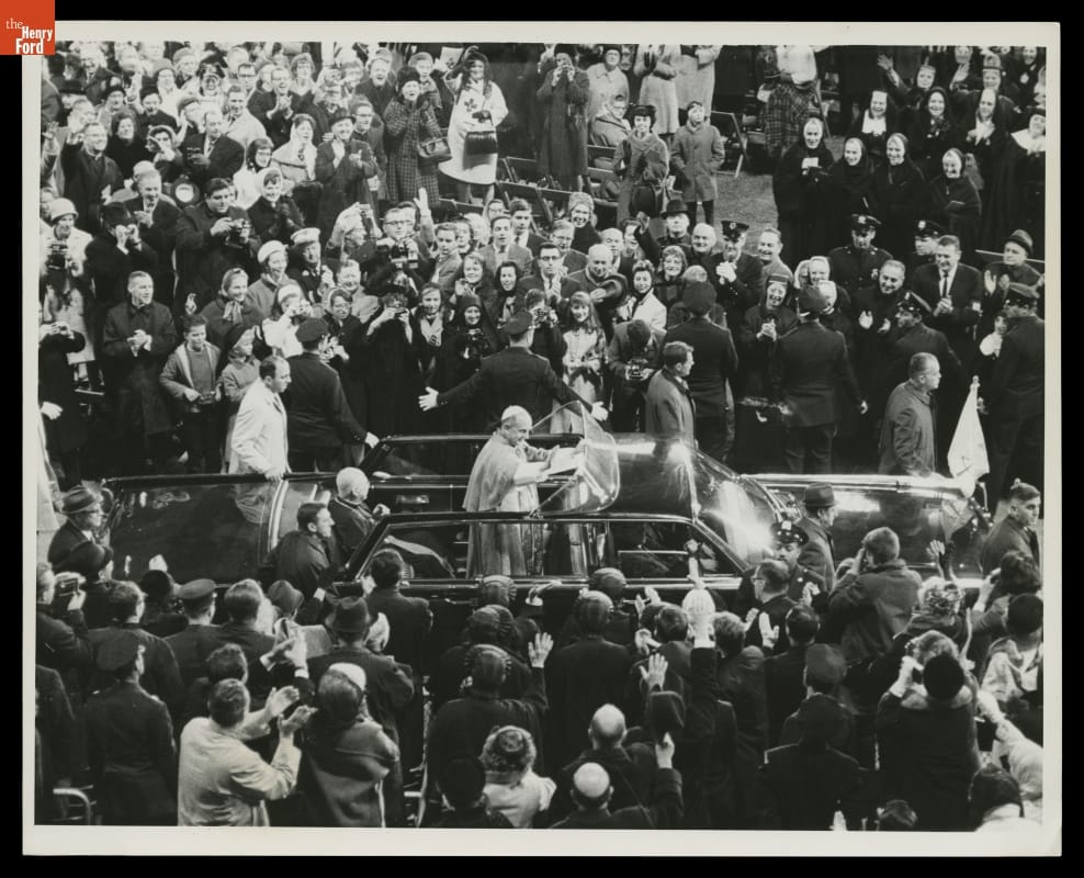 Man in robe and skullcap stands in a limousine, waving, in a dense crowd of people