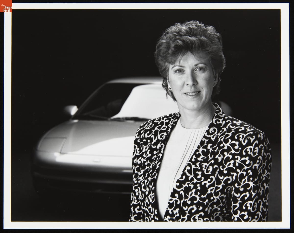 Woman with short hair wearing a jacket with a stylized vine pattern poses in front of a car