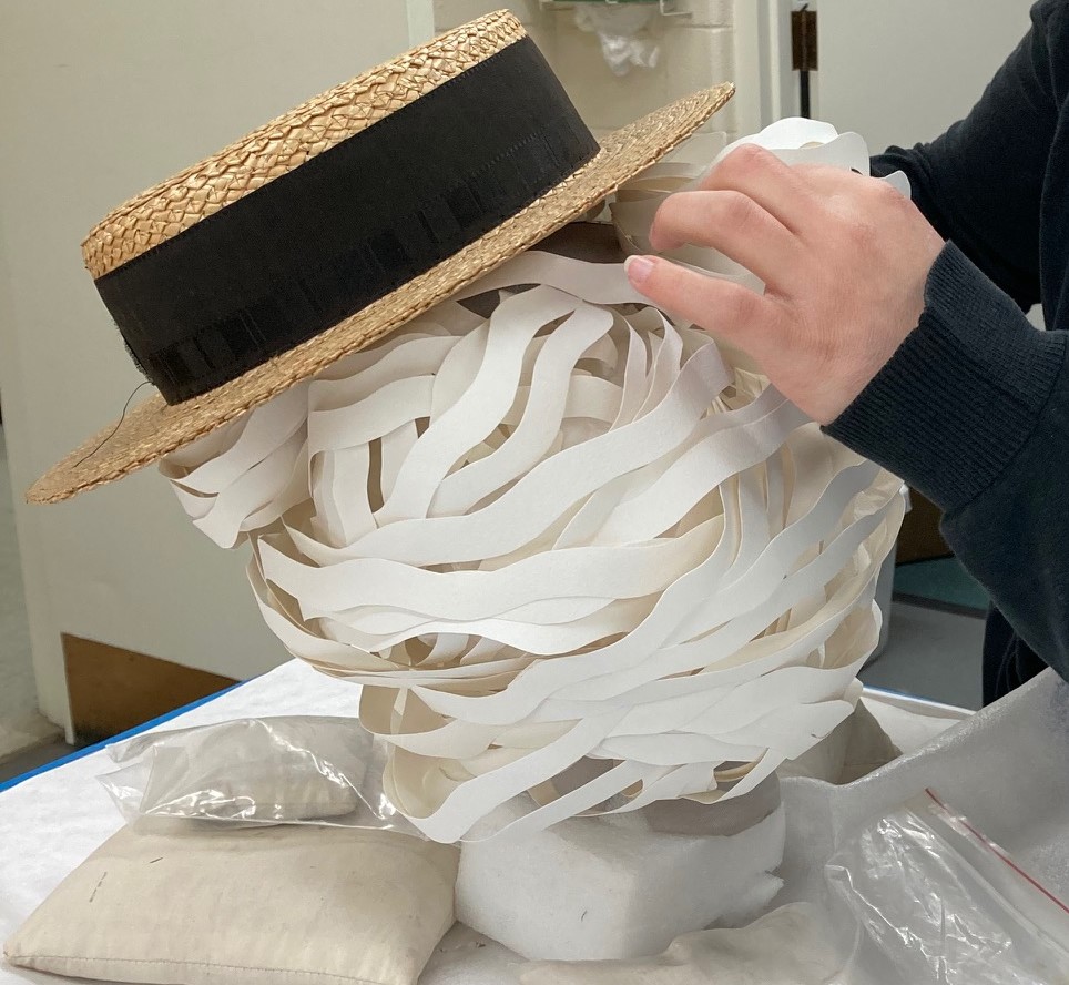 White cut-paper shape with straw boater hat on top is shaped by two hands