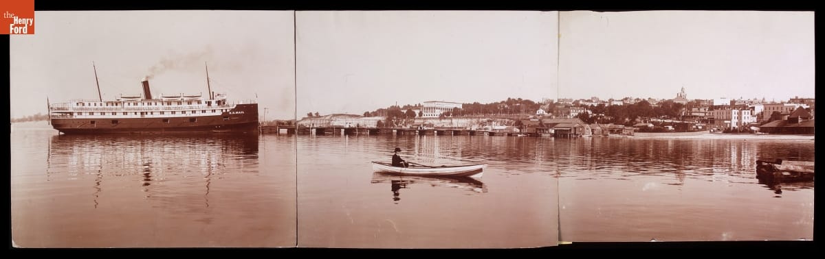 View from the Harbor, Petoskey, Michigan, circa 1906 Man in a rowboat paddles near a larger steamer in a harbor in front of docks and a town