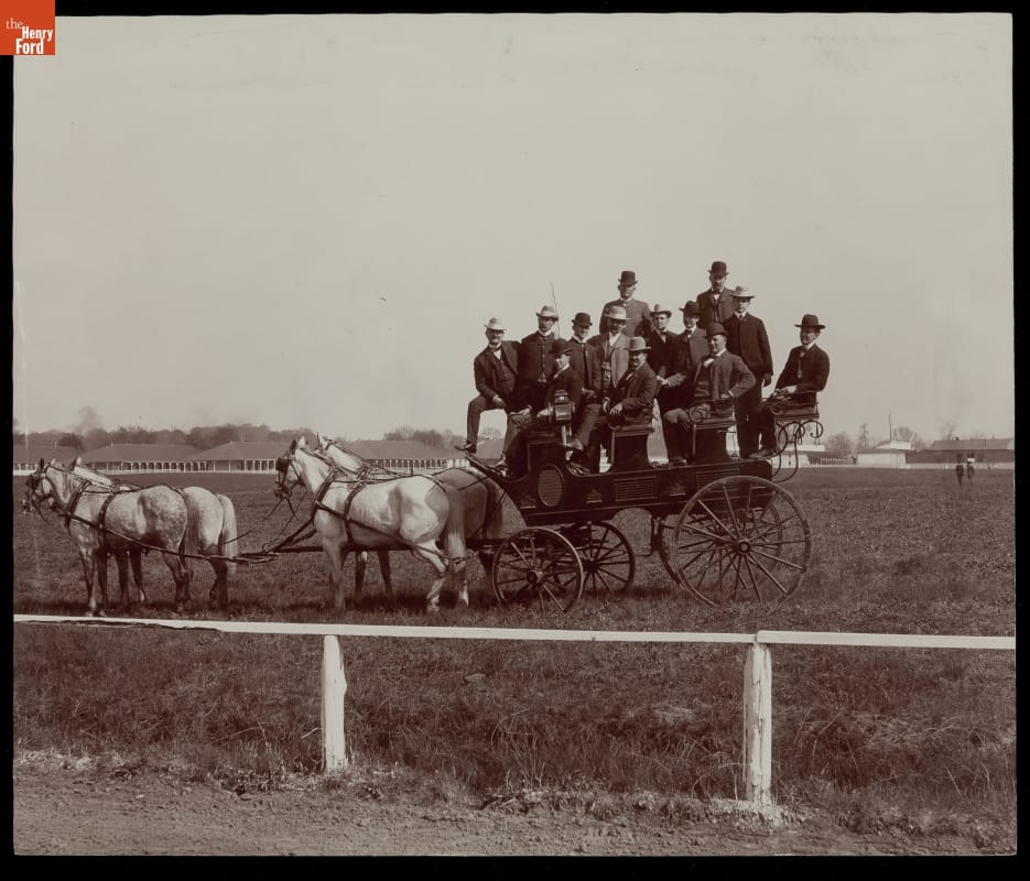 Four horses harnessed to a coach with quite a few passengers seated and standing in it, in a grassy area behind a white fence