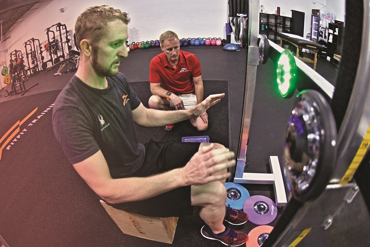 Man in t-shirt and shorts sits in front of lighted device in gym with hands raised as another man kneels next to him