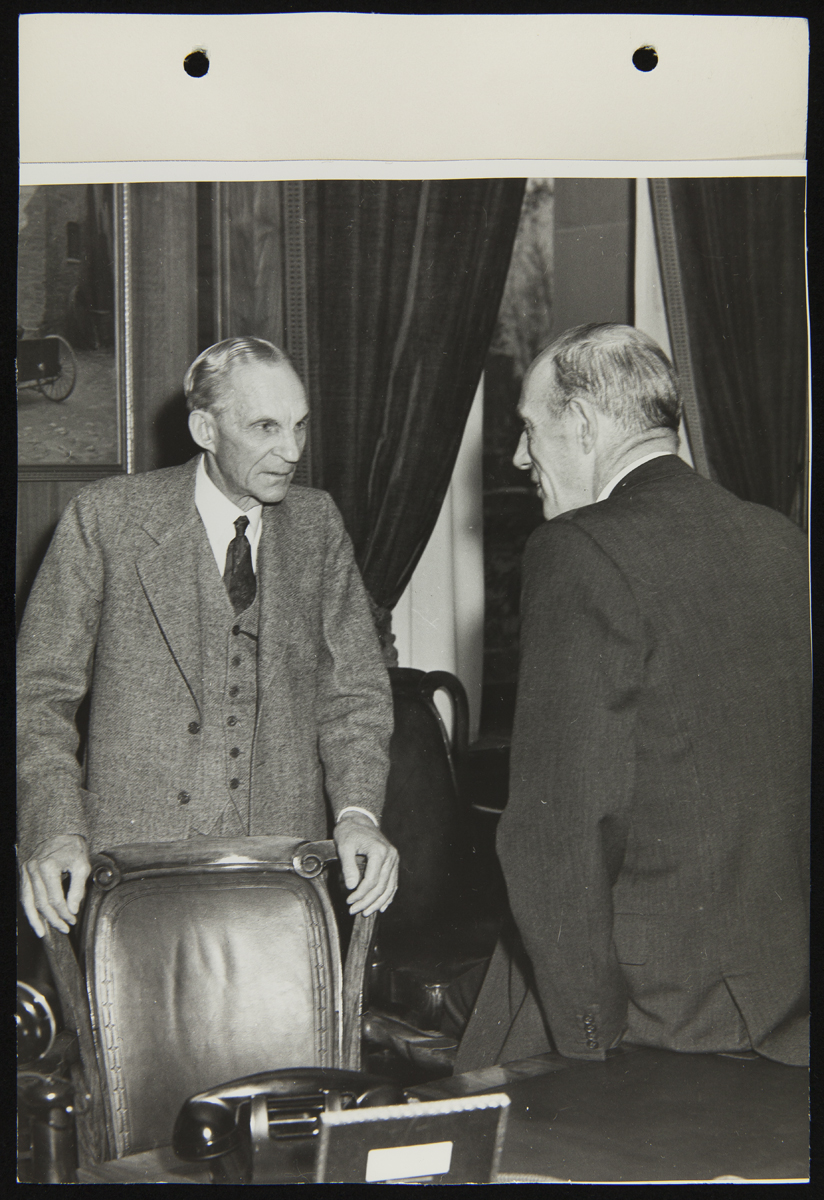 Two men, one holding onto the back of a chair, talk in an office
