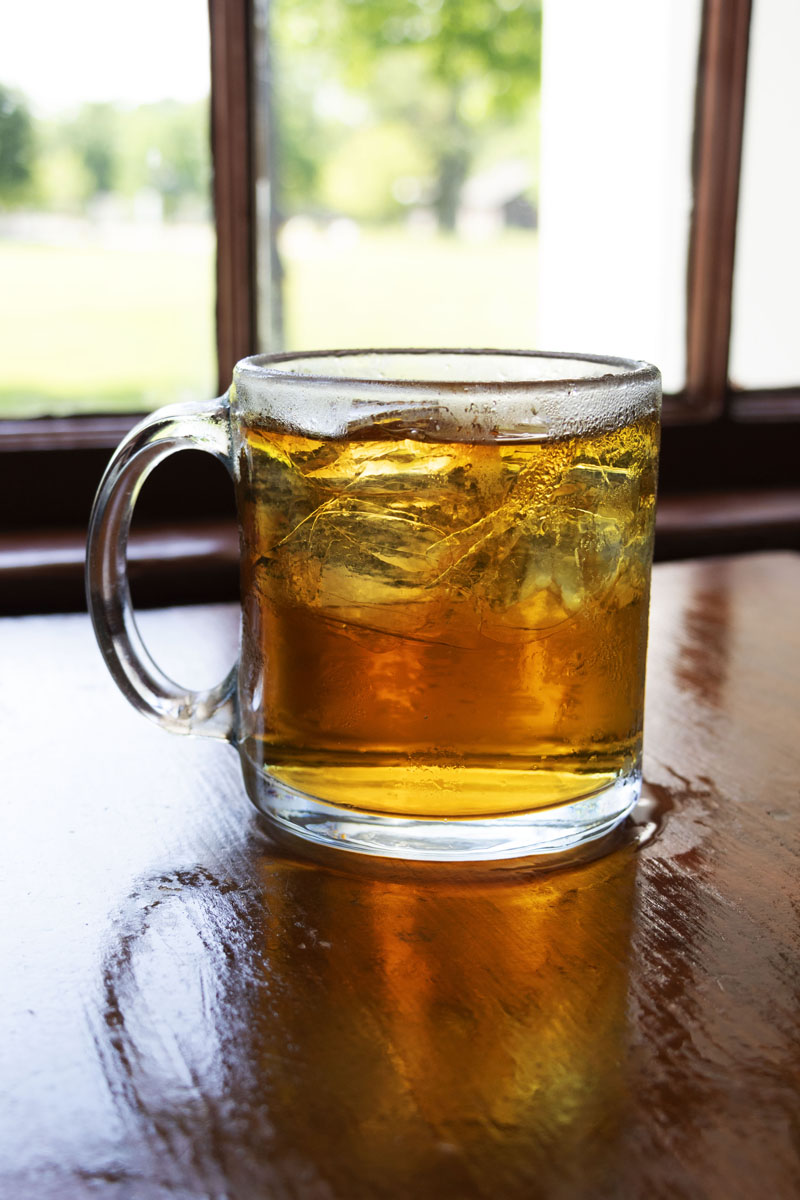 Clear glass mug with amber liquid and ice inside, sitting on wooden table in front of window