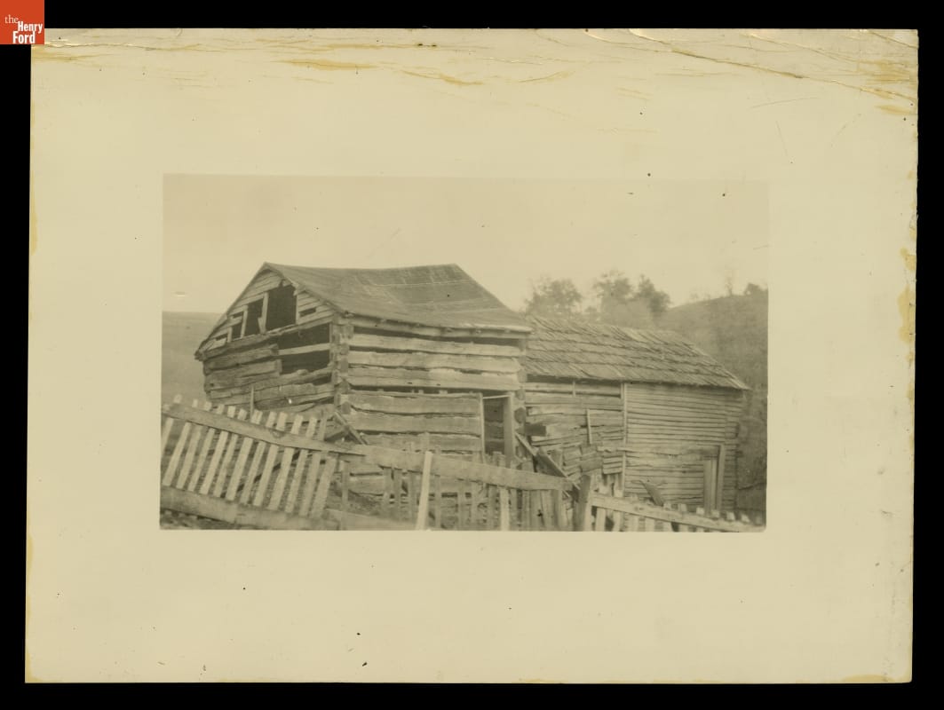 Matted photograph of run-down looking wooden building with equally run-down looking fence in front