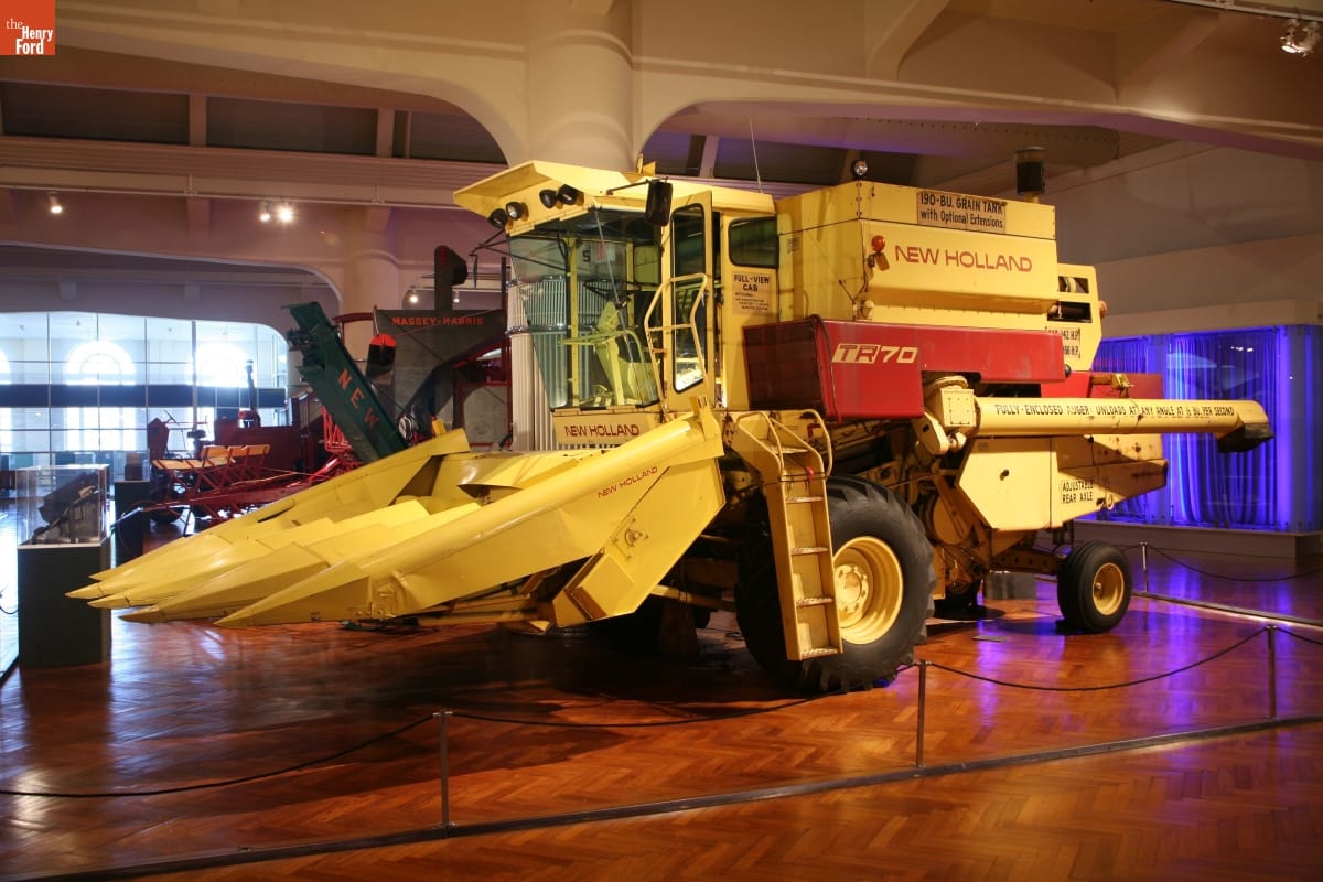 Large yellow piece of agricultural equipment with large tines at front below a cab, in a large space with wooden floor and other agricultural equipment