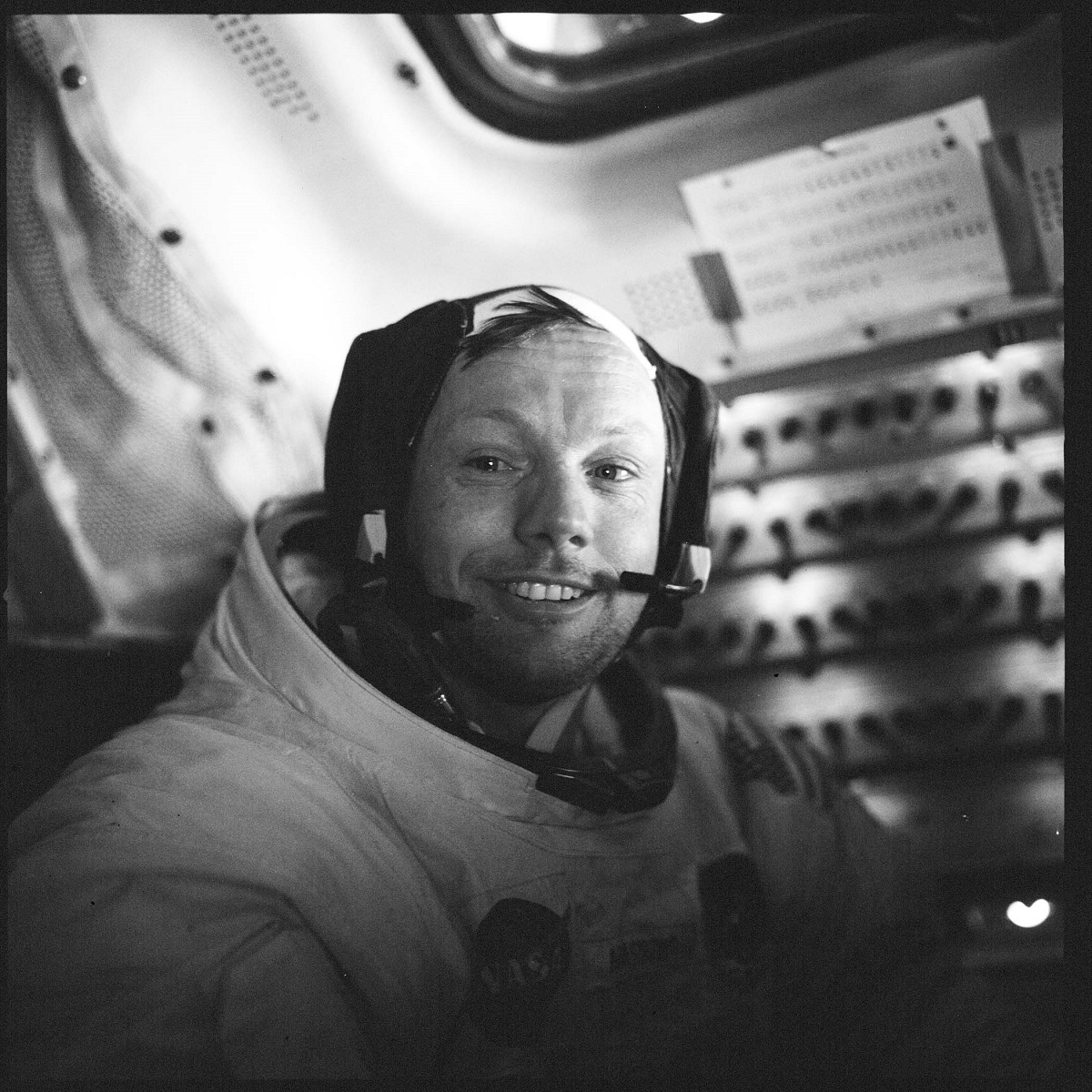 Man in spacesuit smiles at camera with a panel of switches behind him