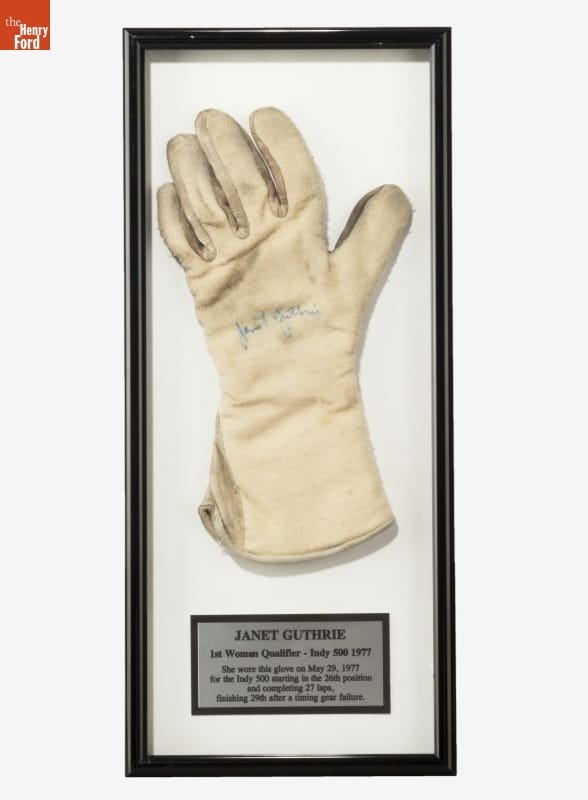 White or cream-colored glove with signature in blue ink in center of palm, mounted under glass in a black frame with a plaque containing text underneath