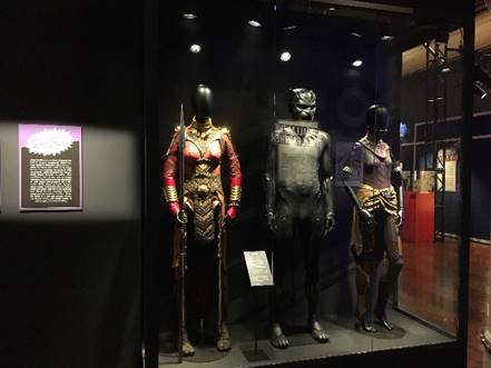 Costumes from the Black Panther film in the "Marvel: Universe of Super Heroes" exhibit Three mannequins in costumes in case with text panel to left