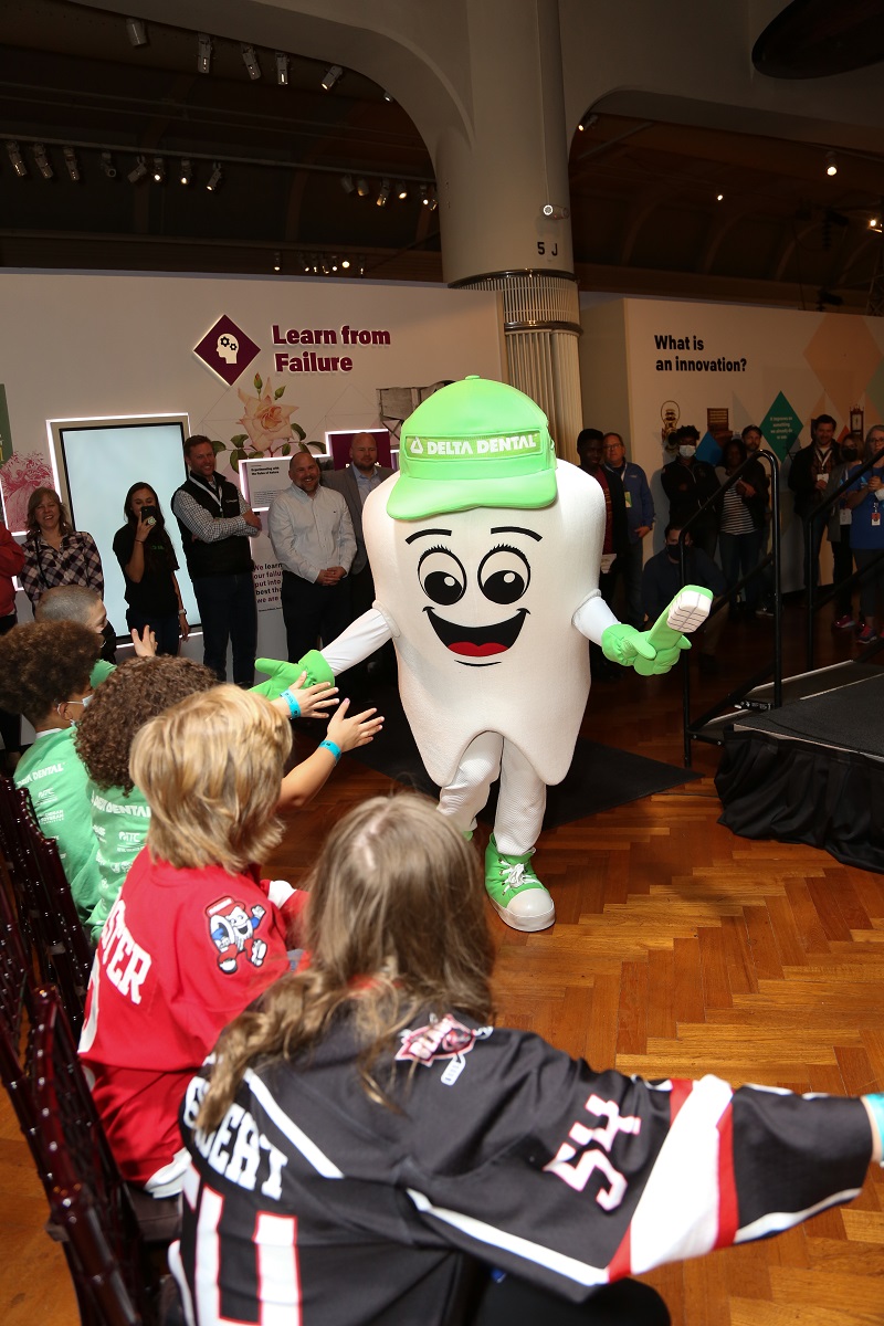 Someone dressed in a tooth costume, with a large smiley face and green hat, gloves, and shoes, entertains a group of people