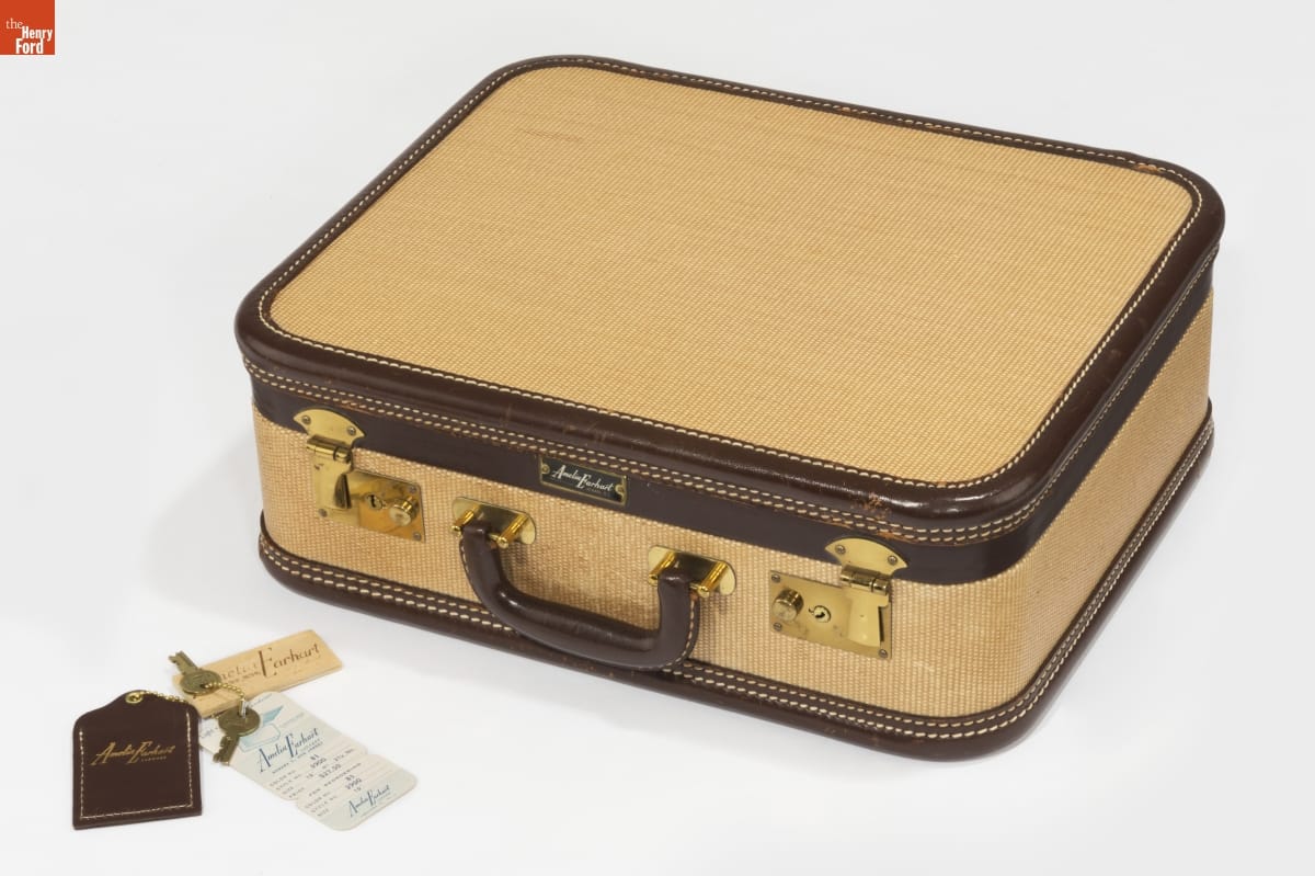 Camel-colored suitcase with brown trim and gold hardware; keys and a label sit nearby
