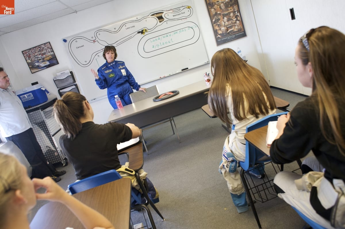 Woman in blue jumpsuit stands behind a table, in front of a whiteboard, at the front of a room with young women sitting in school desks 