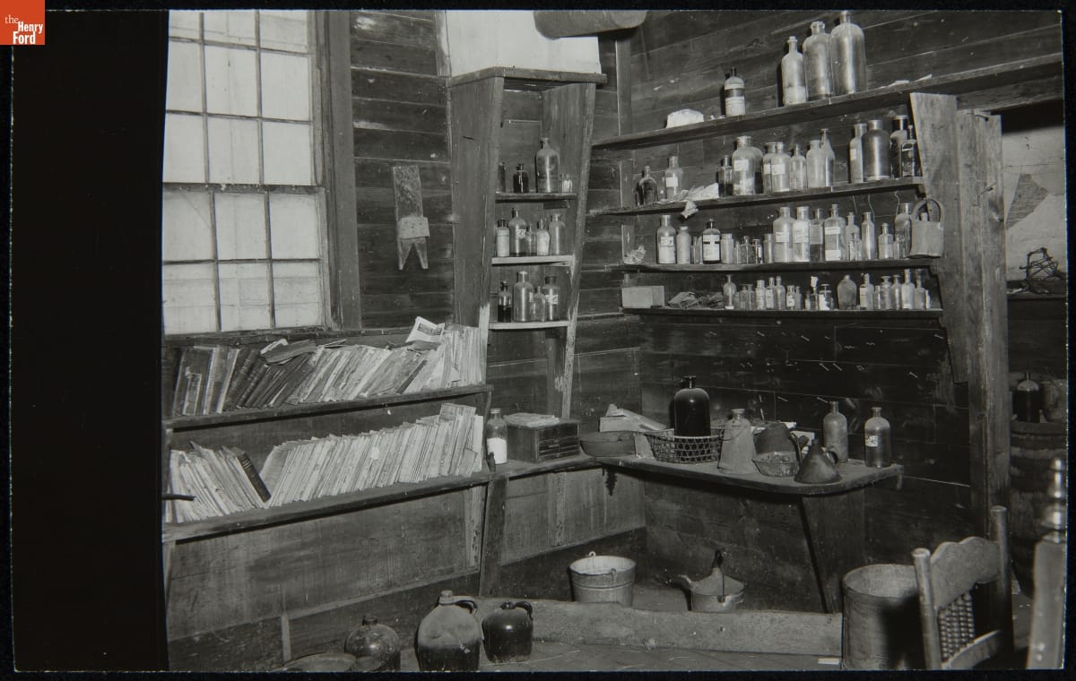 Black-and-white photo of room interior with wooden walls lined with wooden shelves containing books, bottles, and jars