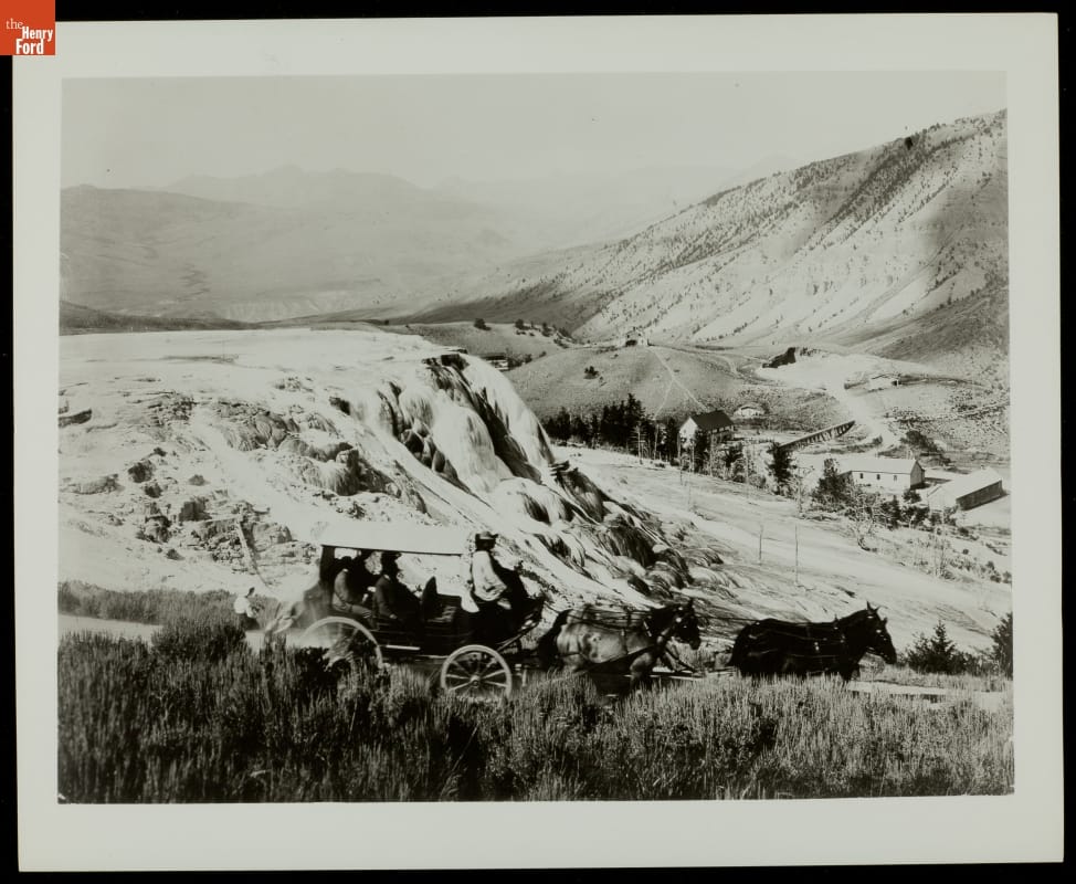 Black-and-white photo of horse-drawn carriage driving past a large rock formation among steep hills and valleys