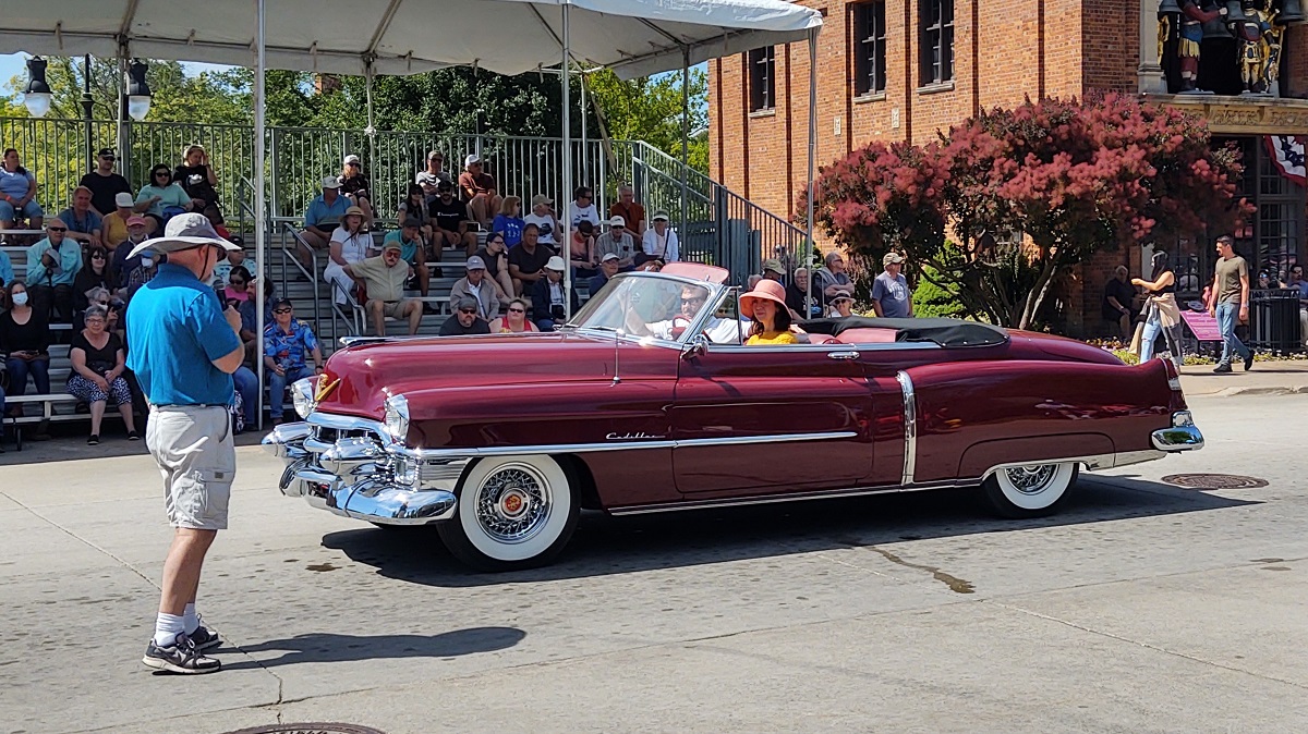 A long, burgundy-colored convertible with dramatic lines drives past a man with a microphone in front of grandstands with seated spectators