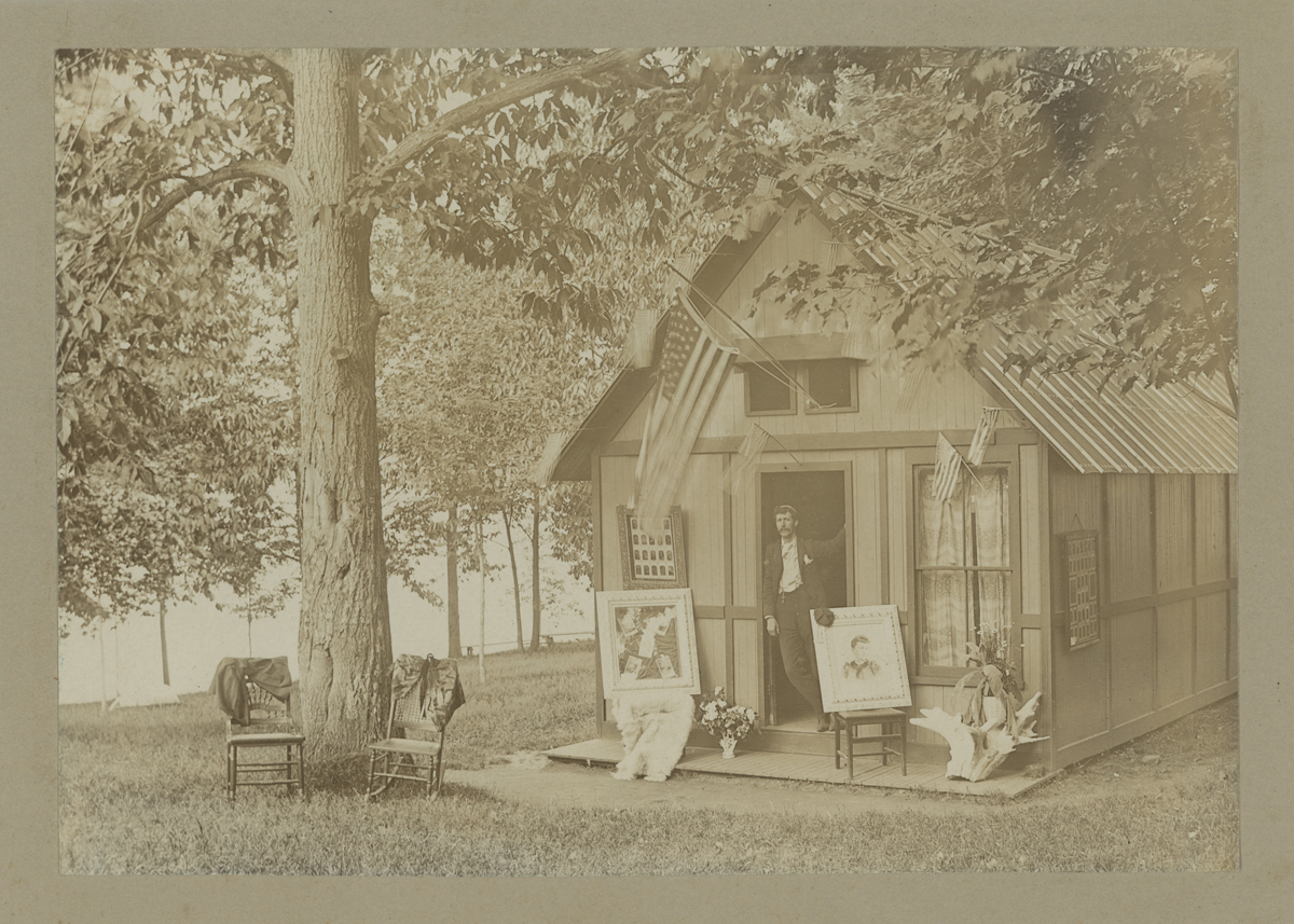 Man leaning in doorway of small wooden building in wooded location; chairs and portraits outside