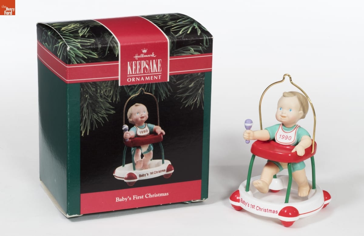 Ornament with a baby or toddler in a walker with "Baby's 1st Christmas" written on it and "1990" on a bib around the baby's neck; it sits next to a green and red box with an image of the same ornament on it