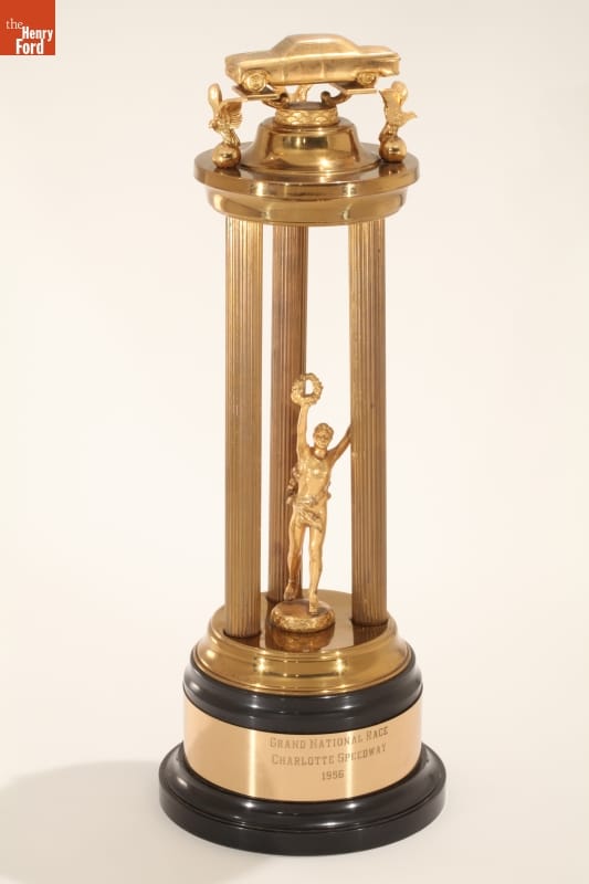 NASCAR Grand National Racing Trophy Won by Buck Baker, 1956 Gold trophy with three tall pillars with car on top; text on black and gold base