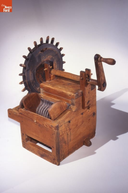 Wooden box-like machine with toothed wheel on one side and handle on the other